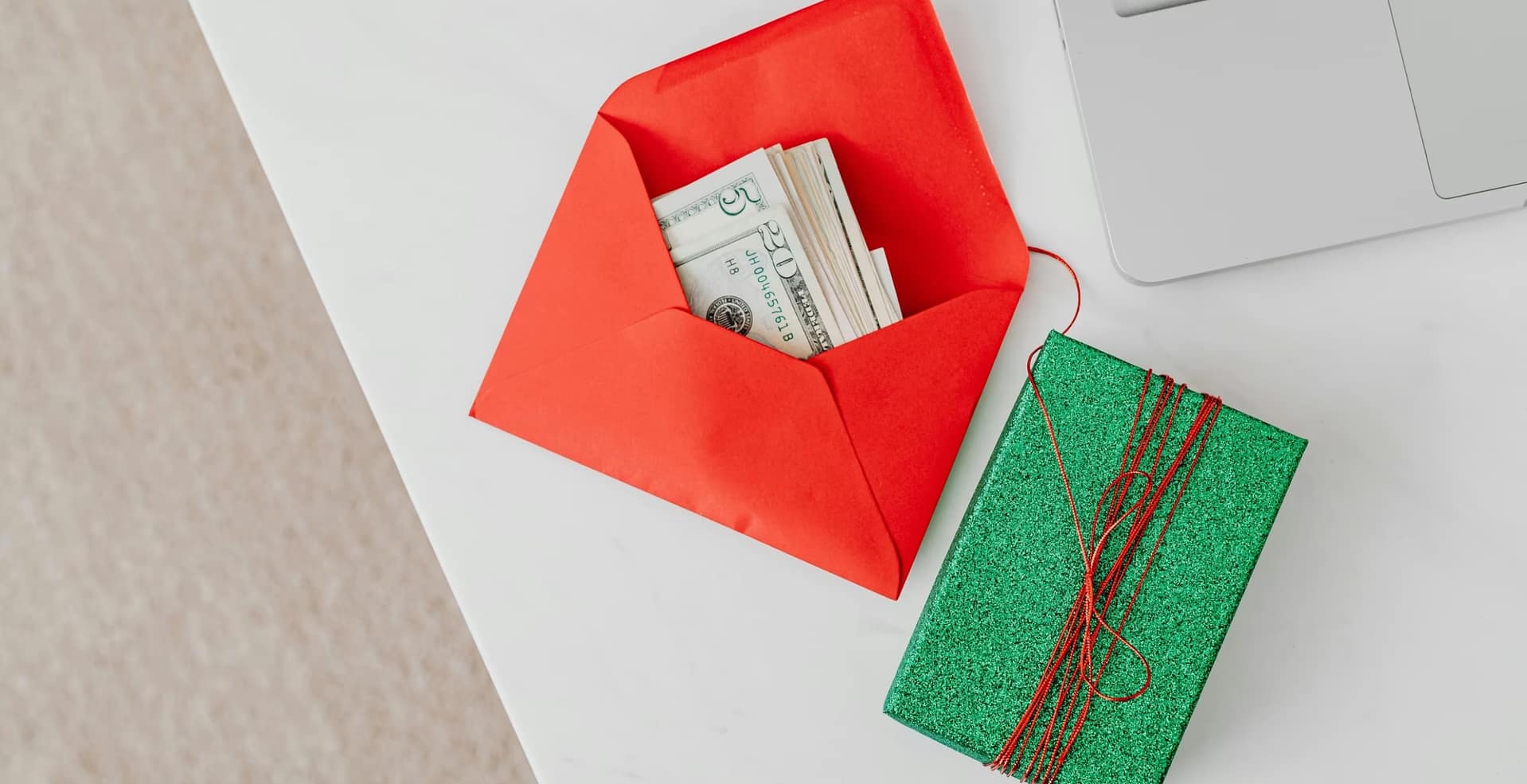 A green gift box and red envelope with cash on a desk beside a laptop, emphasizing holiday or financial themes.